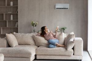 Femme allongée sur son sofa, sur le dos, utilisant son téléphone portable au-dessus d'elle, en dessous de sa thermopompe.